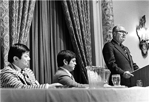 Three men sit at a table facing a man standing at a podium. Decorative drapes hang behind them. 